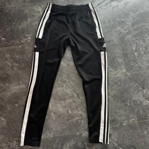 Svarta Adidas träningsbyxor - Svarta träningsbyxor från Adidas med klassiska vita ränder längs sidorna och Adidas-logga på låret. 