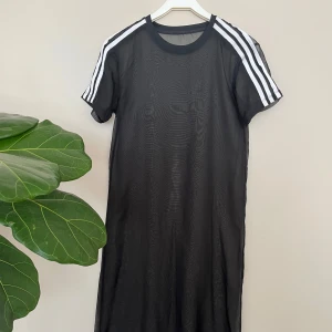 Svart klänning från Adidas - Svart lång meshklänning med enkel följsam stretchig underklänning från Adidas. Klassiska vita ränder på axlarna och korta ärmar. Klänningen har rund halsringning och Adidas-logga på bröstet. Snyggt både var för sig eller ihop.
