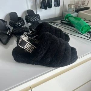 Svarta fluffiga sandaler från UGG - Säljer ett par svarta fluffiga sandaler från UGG med breda band och elastisk rem med UGG-logga. Perfekta för att mysa runt hemma eller som statement piece till din outfit.