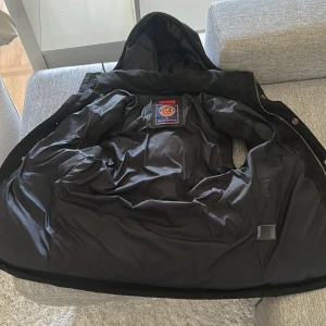 Svart pufferväst med huva från Superdry - Säljer en svart pufferväst från Superdry med huva och dragsko. Västen har två fickor med dragkedja och knäppning framtill. Perfekt för lager-på-lager och håller dig varm under kyliga dagar.