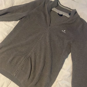 Grå ziptröja från Gant - Säljer en stilren grå ziptröja från Gant med diskret logga på bröstet. Tröjan har lång ärm, ribbade muddar och dragkedja hela vägen. Perfekt för lager-på-lager och enkel att matcha med det mesta.