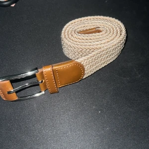Beige flätat bälte med bruna skinn-detaljer - Säljer ett stilrent beige bälte med flätad design och bruna skinn-detaljer vid spännet och ändarna. Bältet har ett silvrigt metallspänne och passar perfekt till jeans eller chinos. Enkel och snygg accessoar för att lyfta din outfit.