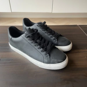 Helt nya oanvända svarta sneakers - Säljer ett par stilrena svarta sneakers med svart snörning och vit platt sula. Skorna har en klassisk design med rund tå och passar perfekt till en avslappnad look.