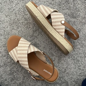 Beige sandaler med remmar från Graceland - Snygga sandaler i beige med breda, korslagda remmar och justerbar hälrem. Skorna har en platt sula med flätad detalj runt kanten och ett diskret randigt mönster på remmarna. Perfekta för varma dagar.