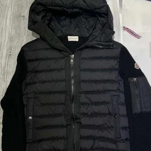 Svart pufferjacka med stickade ärmar från Moncler - Säljer en svart pufferjacka från Moncler med stickade ärmar och huva. Jackan har dragkedja framtill, två fickor med knappar och en extra ficka på ärmen. Perfekt för kyliga dagar och riktigt snygg streetstil.