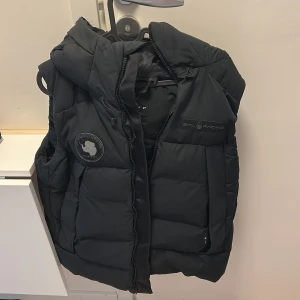 Svart dunväst med huva från Sail Racing - Säljer en svart dunväst från Sail Racing med huva och dragkedja. Västen har en broderad logotyp på bröstet och en rund patch på sidan. Perfekt för lager-på-lager under kyliga dagar.