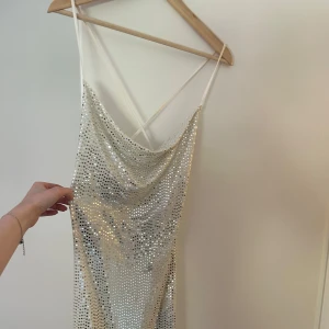 Glittrig silver klänning - Supersnygg klänning med korsad rygg och draperad urringning. Klänningen har en figurnära passform och är perfekt för att sticka ut på en fest eller på klubben. Längden är relativt kort och den är lite genomskinlig så rekomenderar hudfärgade underkläder