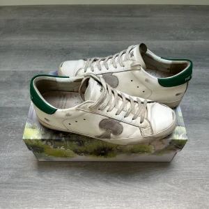 Vita sneakers från The Gallerist med gröna detaljer - Säljer ett par vita sneakers från The Gallerist med gröna hälpartier och ett stort svart klövertryck på sidan. Skorna har snörning och platt sula, perfekt för en avslappnad stil. Levereras i originalkartong med konstnärligt motiv. 