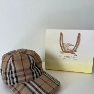 Burbbery keps med monogram - Säljer en klassisk beige rutig keps från Burberry med det ikoniska rutmönstret i svart, rött och vitt. Kepsen har böjd skärm och justerbar passform. Perfekt accessoar för en stilren look.