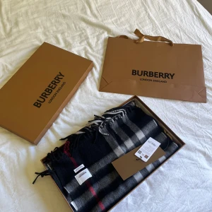Rutig halsduk från Burberry - Säljer en klassisk rutig halsduk från Burberry i mörka färger med inslag av rött, vitt och grått. Halsduken är elegant och levereras i originalförpackning med tillhörande påse. Perfekt accessoar för att lyfta din outfit.