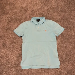 Ljusblå pikétröja från Polo Ralph Lauren - Säljer en klassisk ljusblå pikétröja från Polo Ralph Lauren med korta ärmar och krage. Tröjan har den ikoniska broderade loggan i orange på bröstet och knäppning vid halsen. Perfekt för en stilren och avslappnad look.