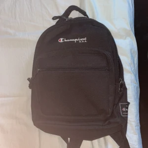 Svart ryggsäck från Champions - Säljer en svart ryggsäck från Champions med broderad logga framtill. Väskan har ett stort huvudfack, ett mindre ytterfack med dragkedja och justerbara axelband. Perfekt för skola eller träning. Enkel och stilren design.