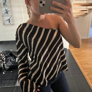 Svart och vit randig oneshoulder blus - Säljer en svart och vit randig blus med oneshoulder-design. Blusen har långa ärmar och ett luftigt, avslappnat snitt. Perfekt för dig som vill sticka ut med en trendig och stilren topp.
