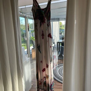 Färgglad långklänning från Natthanicha Fashion - Säljer en långklänning med färgstarkt blommigt mönster och djup v-ringning. Klänningen har breda axelband och är i tunnt, mjukt material. Perfekt för sommarens varma dagar.