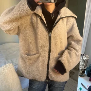 Beige teddyjacka med dragkedja - Mysig beige teddyjacka med dragkedja framtill och stora fickor. Jackan har lång ärm och en avslappnad passform, perfekt för kyliga dagar. Enkel och stilren design som passar till många outfits.