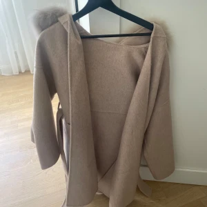 Beige kappa med pälskrage och bälte - Säljer en beige kappa med stor pälskrage och bälte i midjan. Jackan har lång ärm och en mjuk, ullig känsla. Perfekt för kyliga dagar när du vill vara både varm och snygg. I storlek S använt fåtal gånger.