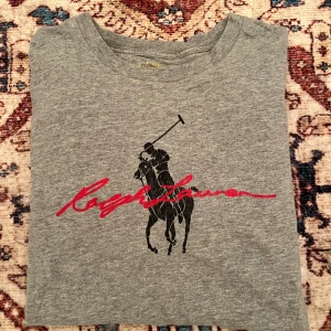 Grå t-shirt från Polo Ralph Lauren - Grå t-shirt från Polo Ralph Lauren med klassiskt tryck av en polospelare och röd signaturtext på bröstet. Rund halsringning och mjukt material som känns skönt mot huden. Perfekt för en avslappnad och stilren look.