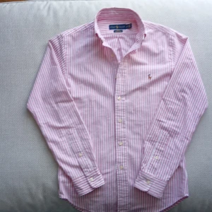 Rosa och vit randig skjorta från Ralph Lauren - Säljer en slim fit skjorta från Ralph Lauren i rosa och vitt randigt mönster. Skjortan har långa ärmar, klassisk krage och knappar framtill. Broderad logga på bröstet. Perfekt för dig som gillar stilrena plagg med snygga detaljer.