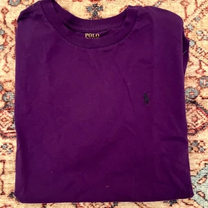 Lila t-shirt från Polo Ralph Lauren - Säljer en lila t-shirt från Polo Ralph Lauren med klassisk broderad logga på bröstet. T-shirten har rund halsringning och är tillverkad i mjuk bomull. Perfekt för en stilren och avslappnad look.