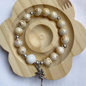 Armband med vita pärlor  - Snyggt armband med runda vita pärlor och silverfärgade kulor. Armbandet har en söt ängelberlock som hänger ner. Perfekt accessoar för att ge din outfit en unik touch.