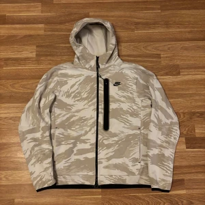 Nike beige camo tech fleece - Säljer en snygg beige och vit camouflage vindjacka från Nike med huva och svart dragkedja. Jackan har en bröstficka med vertikal dragkedja och Nike-logga på bröstet. Perfekt för dig som gillar streetwear och sportig stil. Nypris 1,499kr. Kan lämnas i Kristianstad, Lund och Malmö.