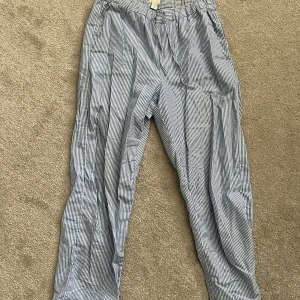 Blåvitrandiga byxor med snörning - Säljer ett par blåvitrandiga byxor från ARKET med snörning i midjan. Byxorna har en avslappnad passform och är perfekta för varmare dagar. Tunt och skönt material som känns luftigt och bekvämt.