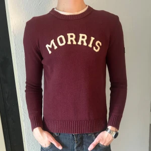 Vinröd stickad tröja från Morris - Vinröd stickad tröja från Morris med vit broderad logga på bröstet och patch med brittisk flagga på ärmen. Rund halsringning och ribbade muddar vid ärmslut och nederkant. Perfekt för en stilren och klassisk look.
