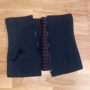 Svart midjebälte med hysk- och hakstängning - Säljer ett svart midjebälte med flera rader hyskor och dragkedja framtill. Bältet är format för att sitta tajt runt midjan och passar perfekt för att skapa en markerad siluett. Materialet är stretchigt och följsamt. Säljer då den är för stor🥲