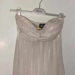 Beige axelbandslös långklänning från Nelly - Elegant beige långklänning från Nelly med glittrig, draperad överdel och axelbandslös design. Klänningen har ett vackert fall och passar perfekt till speciella tillfällen. Mjuk och lätt i materialet för en lyxig känsla.