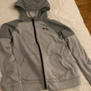 Ljusgrå hoodiekofta från Under Armour - Snygg ljusgrå kofta från Under Armour med svart dragkedja och logga på bröstet. Koftan har huva och två stora fickor framtill. Perfekt för träning eller chill.