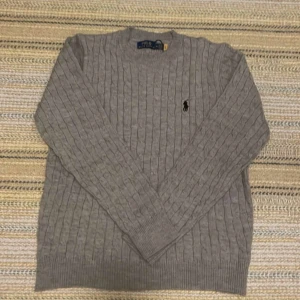 Ralph lauren ull tröja  - Säljer en klassisk grå kabelstickad tröja från Polo Ralph Lauren med broderad logga på bröstet. Tröjan har rund halsringning och långa ärmar, perfekt för lager-på-lager under kyligare dagar.