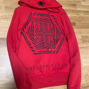 Röd hoodie från Philipp Plein - Säljer en röd hoodie från Philipp Plein med svart grafiskt mönster på bröstet och logotyptryck nedtill. Tröjan har huva med dragsko och en metallbadge på huvan. Säljes med originallåda.