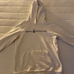 Vit hoodie från Sail Racing - Vit hoodie från Sail Racing med svart logga på bröstet. Klassisk känguruficka framtill och huva. Perfekt för en avslappnad stil. Priset kan diskuteras. Ny pris 1200.