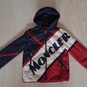 Moncler tunn dunjacka med huva stl 164 - Snygg dunjacka från Moncler med stor logga över bröstet. Jackan har huva, dragkedja och två fickor framtill. Färgblock i marinblått, rött och vitt med broderad Moncler-patch på ärmen. Perfekt för dig som vill sticka ut. Liten fläck ovanför bokstaven R men syns knappt. 