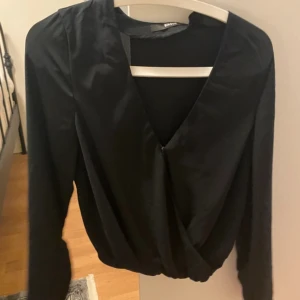 Svart omlottblus från BikBok - Säljer en svart omlottblus från BikBok i storlek S. Blusen har lång ärm och djup v-ringning framtill. Perfekt för dig som gillar stilrena och enkla plagg med en twist.