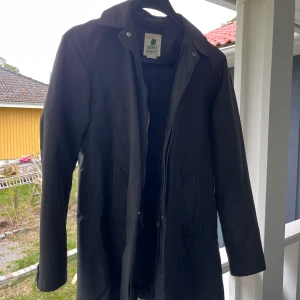 Svart trenchcoat  från Sierra Designs - Säljer en stilren svart trench coat från Sierra Designs. Jackan har klassisk krage, dold dragkedja och knäppning framtill samt två snedställda fickor. Perfekt för dig som vill ha en enkel och tidlös look.