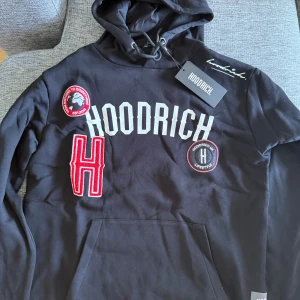 Svart hoodie från Hoodrich med broderade detaljer - Säljer en svart hoodie från Hoodrich med broderade märken och stor röd H på framsidan. Tröjan har huva med dragsko och känguruficka. Perfekt för dig som gillar streetwear och vill sticka ut med snygga detaljer.