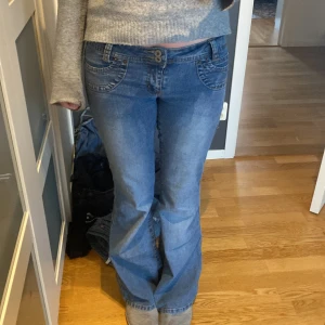 Blå bootcut jeans med låg midja - Säljer ett par klassiska blå bootcut jeans med låg midja och snygga detaljer på fickorna. Jeansen har en lätt tvättad look och sitter tight upptill med utsvängda ben. Perfekta för en avslappnad och trendig stil.