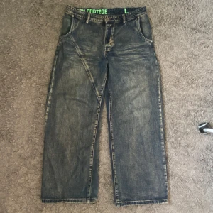 Baggy Jeans från Protemoa - Tunga Jeans av Protemoa/Non Protégé i storlek L.  Riktig kvalité och många fina detaljer. Jeansen är lite mörkare och blåare än vad de ser ut på bilderna. Ända defekten är heel drag tydlig på sista bilden. DMa om de finns frågor eller om mått. Passar bra som W33-W35