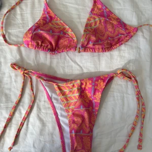 Färgglad bikini med blommönster - Snygg och färgglad bikini med blommönster i rosa och orange. Toppen har trekantsform och knytband i nacken och ryggen. Bikinitrosan har knytband på sidorna för justerbar passform. Perfekt för sommarens stranddagar! Ny skick Stl xs 