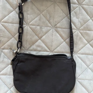 Svart axelväska med kedjedetalj - Snygg svart axelväska med en unik kedjedetalj på axelremmen. Väskan har en dragkedja upptill för säker förvaring av dina saker. Perfekt för att ge en stilren touch till din outfit.