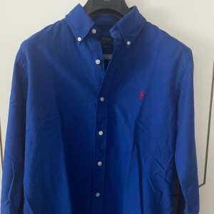 Blå skjorta från Ralph Lauren - Säljer en klassisk blå skjorta från Ralph Lauren med lång ärm och knappar. Skjortan har en broderad logotyp i rött på bröstet och är tillverkad i bomull. Perfekt för en stilren look. Nypris ca 1800kr. Köpt på en av Ralph Laurens officiella outletbutiker, därav prislappen på bilden. Riktigt snygg, perfekt skjorta till sommaren eller våren. Den är helt ny och oanvänd med alla lappar kvar. 