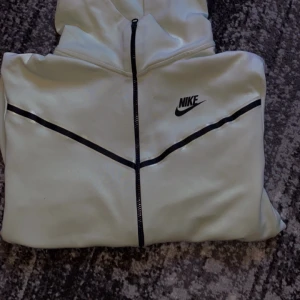 Vit huvtröja från Nike - Säljer en stilren vit huvtröja från Nike med svarta detaljer och dragkedja. Tröjan har en klassisk Nike-logga på bröstet och en bekväm passform. Perfekt för en sportig look eller avslappnade dagar.