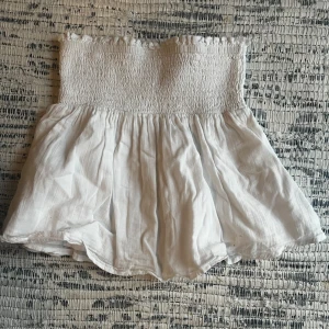 Vit kjol/topp från Gina Tricot - Söt vit kjol/topp från Gina Tricot med smockdetalj. Toppen har en kort längd och är gjord i ett lätt material. Om man vill kan man använda den som kjol också. Har en liten fläck men är nästan inte synlig. 