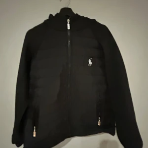 Ralph lauren cardigan  - Snygg svart jacka från Polo Ralph Lauren med dragkedja och broderad logga på bröstet. Jackan har en huva och långa ärmar, perfekt för kyligare dagar. Praktiska fickor med dragkedja framtill.