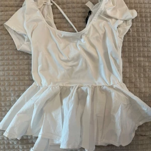 Vit blus från H&M Divided - Säljer en söt vit blus från H&M Divided med volangdetaljer och knytning i ryggen. Blusen har korta ärmar och en luftig passform, perfekt för varma dagar. Den har en rundad halsringning och en lätt, fladdrig nederdel.