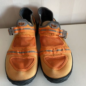 Orange och grå sneakers/sandaler Reebok  - Snygga och unika sneakers från Reebok i en fräsch orange och grå färgkombination. Skorna har ett coolt spänne istället för traditionell snörning och en slitstark sula för bra grepp. Perfekta för dig som vill sticka ut med din stil.