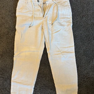 Linnebyxor från Mango - Snygga beige linnebyxor från mango med snörning i midjan och dragkedja. Byxorna har en avslappnad passform och är perfekta för varma dagar. De har två bakfickor med knappar och ett randigt innerfoder.