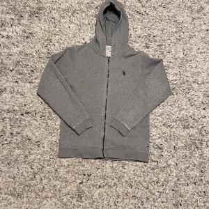 Grå hoodie från U.S. Polo Assn. - Säljer en stilren grå hoodie från U.S. Polo Assn. med dragkedja och klassisk logga på bröstet. Perfekt för en avslappnad stil. Den har långa ärmar och en bekväm huva.