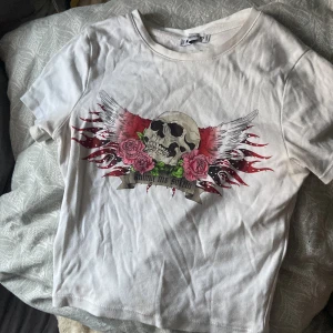 Vit t-shirt med dödskalletryck - Cool vit t-shirt från H&M med ett edgy tryck av en dödskalle, rosor och vingar i rött och svart. Perfekt för en rockig stil. Kortärmad och i mjukt bomullsmaterial.
